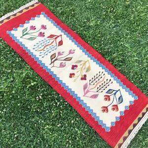May include: A white and red runner rug with a floral pattern. The rug has a red border with a fringe and a white center with a repeating pattern of stylized flowers in shades of red, blue, yellow, and green.