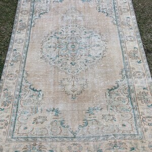 May include: A vintage rug with a light beige background and a faded teal and brown pattern. The rug has a fringed edge.