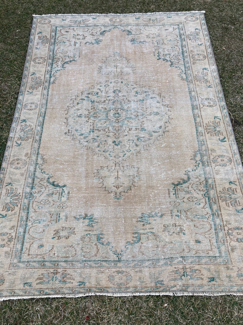 May include: A vintage rug with a light beige background and a faded teal and brown pattern. The rug has a fringed edge.