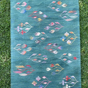 May include: A green rug with a fringe border features a repeating pattern of stylized floral designs in various colors, including pink, blue, yellow, and red.