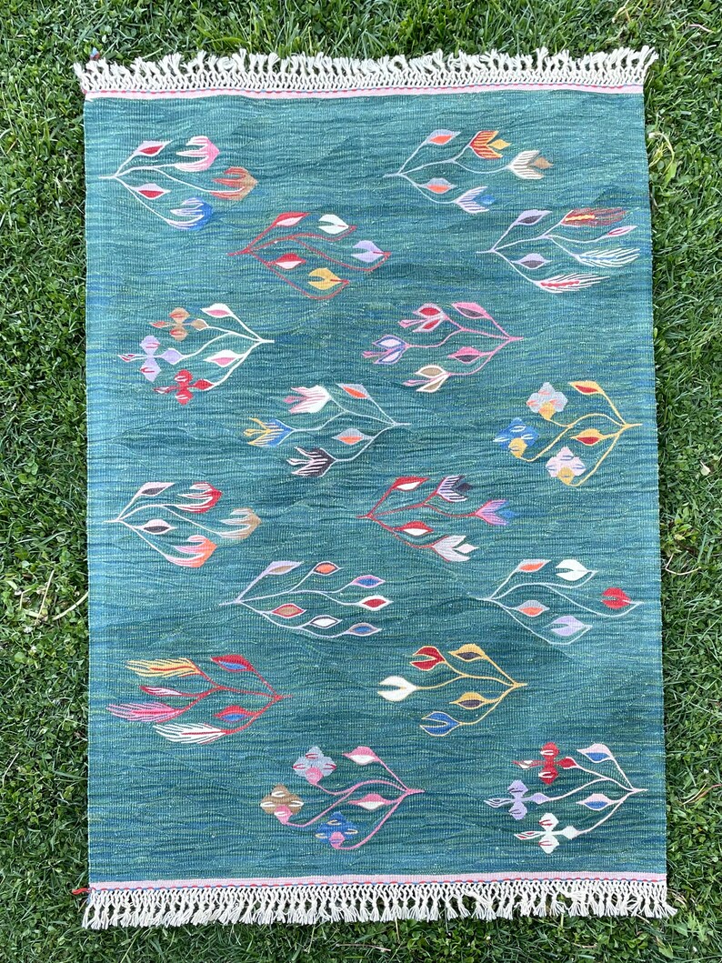 May include: A green rug with a fringe border features a repeating pattern of stylized floral designs in various colors, including pink, blue, yellow, and red.