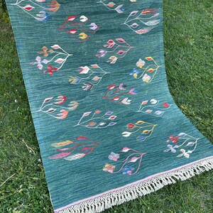 May include: A green rug with a fringe border features a repeating pattern of stylized floral designs in various colors, including pink, red, yellow, blue, and green.