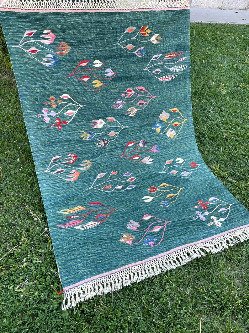 May include: A green rug with a fringe border features a repeating pattern of stylized floral designs in various colors, including pink, red, yellow, blue, and green.
