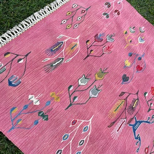 May include: A pink rug with a fringe border features a repeating pattern of stylized floral designs in shades of blue, green, yellow, red, and white.