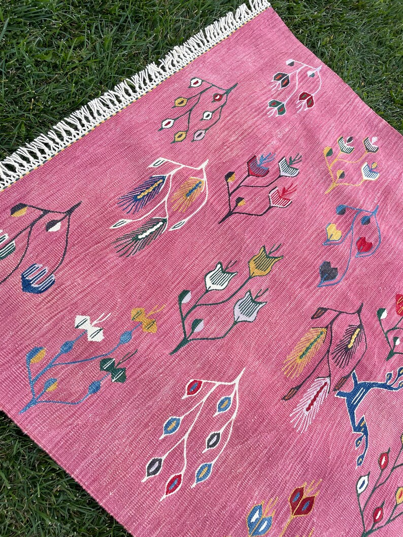 May include: A pink rug with a fringe border features a repeating pattern of stylized floral designs in shades of blue, green, yellow, red, and white.