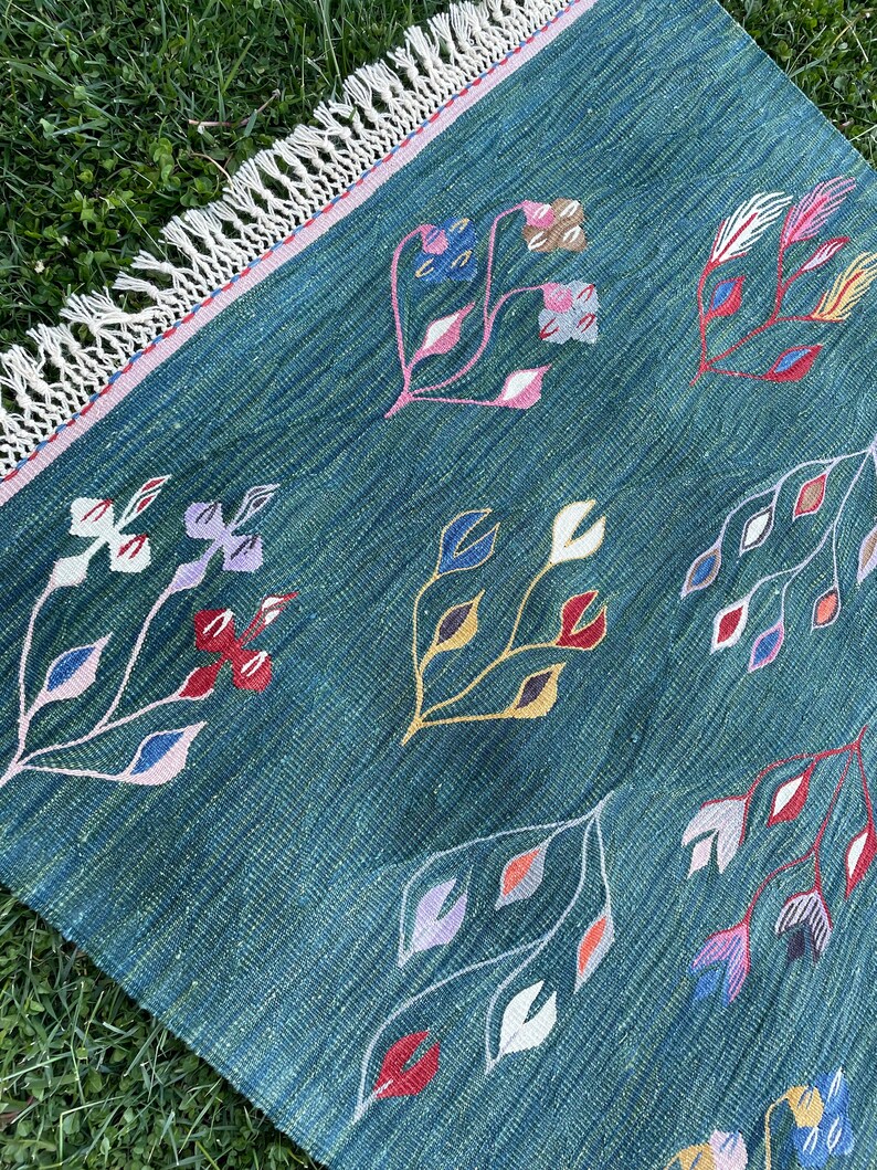 May include: A teal blue rug with a fringe border features a repeating pattern of stylized floral designs in shades of red, pink, yellow, blue, and white.