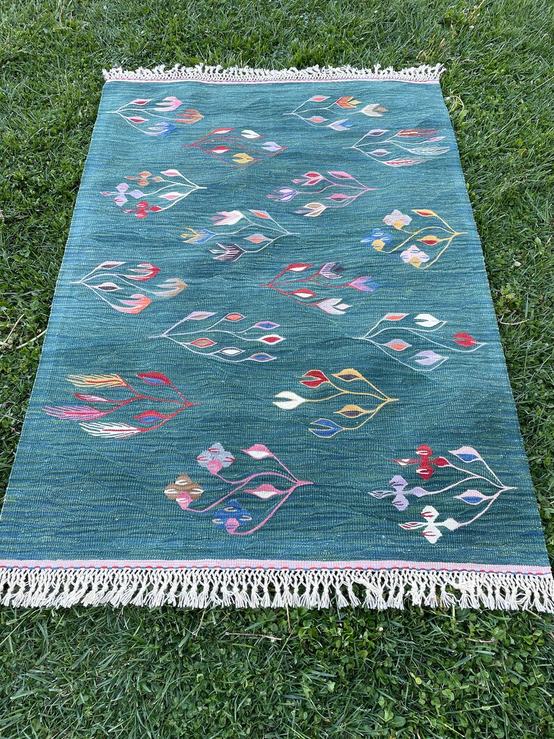 May include: A teal green rug with a fringe border features a repeating pattern of stylized floral designs in shades of pink, red, yellow, blue, and white.
