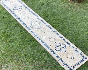 May include: A beige runner rug with a blue and green geometric pattern. The rug has a fringed edge and is laid out on a green grassy surface.