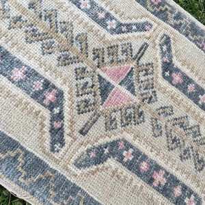 Puede incluir: Una alfombra de color crema con un patr&oacute;n geom&eacute;trico en tonos grises y rosas. La alfombra tiene un borde con flecos y est&aacute; hecha de un material suave y mullido.