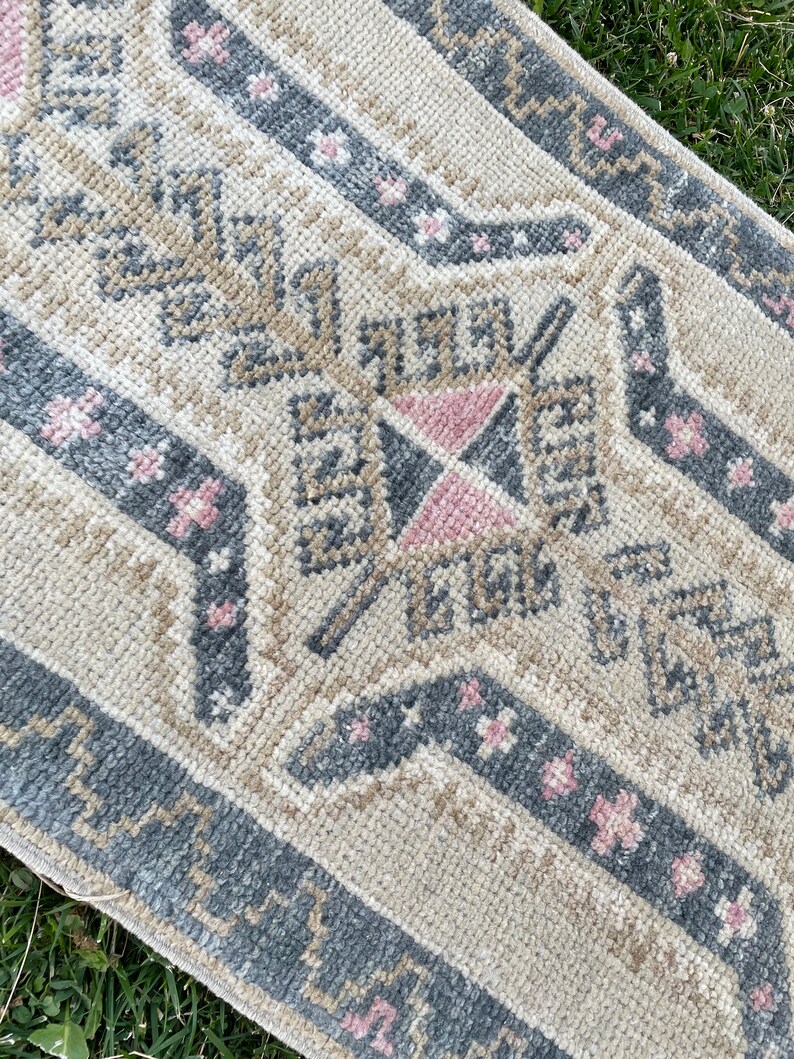 Puede incluir: Una alfombra de color crema con un patr&oacute;n geom&eacute;trico en tonos grises y rosas. La alfombra tiene un borde con flecos y est&aacute; hecha de un material suave y mullido.