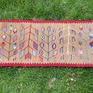 May include: A brown rug with a red border and a pattern of stylized trees with colorful leaves. The rug has a fringe on both ends.