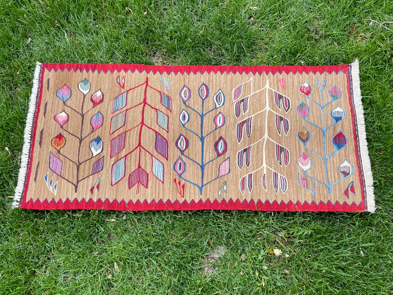 May include: A brown rug with a red border and a pattern of stylized trees with colorful leaves. The rug has a fringe on both ends.