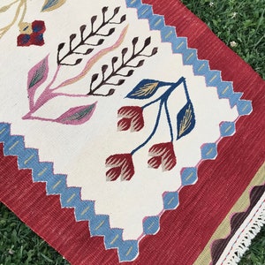 May include: A white and red rug with a floral pattern. The rug has a blue and white border and a fringe on the edges.
