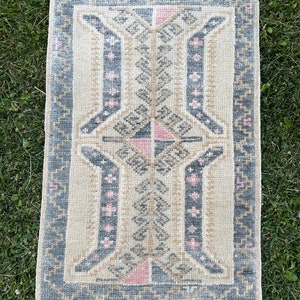 Puede incluir: Una alfombra peque&ntilde;a y rectangular con fondo crema y un patr&oacute;n geom&eacute;trico en tonos grises, rosa y beige. La alfombra tiene un borde de flecos.