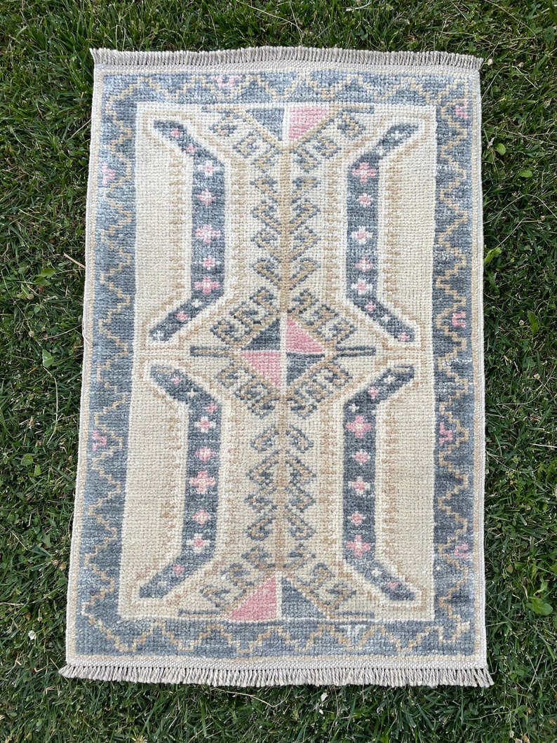 Puede incluir: Una alfombra peque&ntilde;a y rectangular con fondo crema y un patr&oacute;n geom&eacute;trico en tonos grises, rosa y beige. La alfombra tiene un borde de flecos.