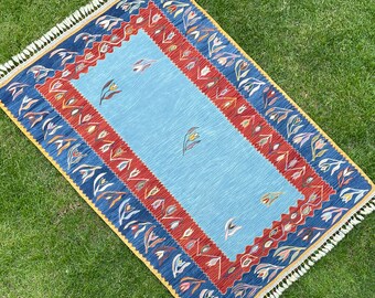 May include: A blue rug with a red, yellow, and white floral pattern. The rug has a fringe border.