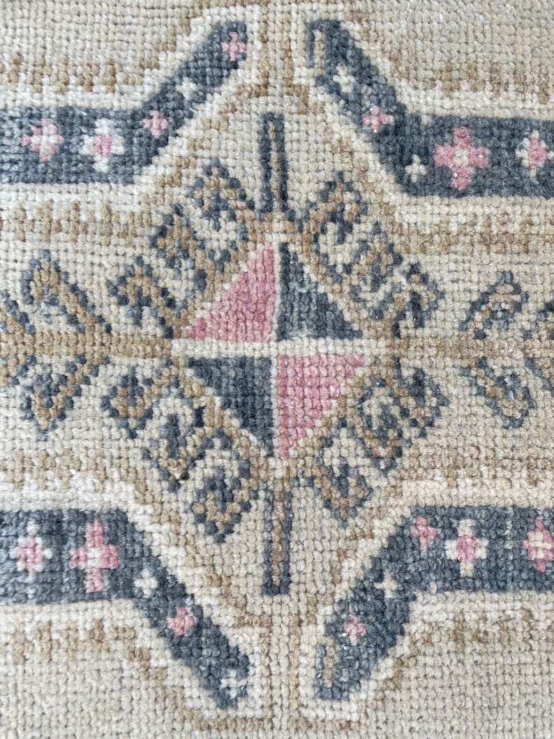 Puede incluir: Primer plano de una alfombra beige con un patr&oacute;n geom&eacute;trico en tonos grises y rosas. El patr&oacute;n presenta una forma de diamante con un diamante m&aacute;s peque&ntilde;o en su interior, rodeado por un borde de diamantes m&aacute;s peque&ntilde;os.