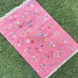 May include: A pink rug with a white fringe border features a colorful floral and geometric pattern. The rug is made of woven fabric and has a traditional design.
