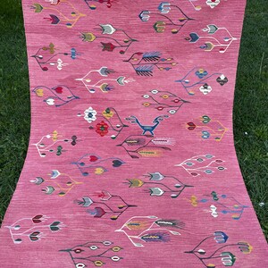 May include: A pink rug with a geometric pattern of stylized flowers and leaves in shades of blue, green, yellow, and red. The rug has a white fringe border.