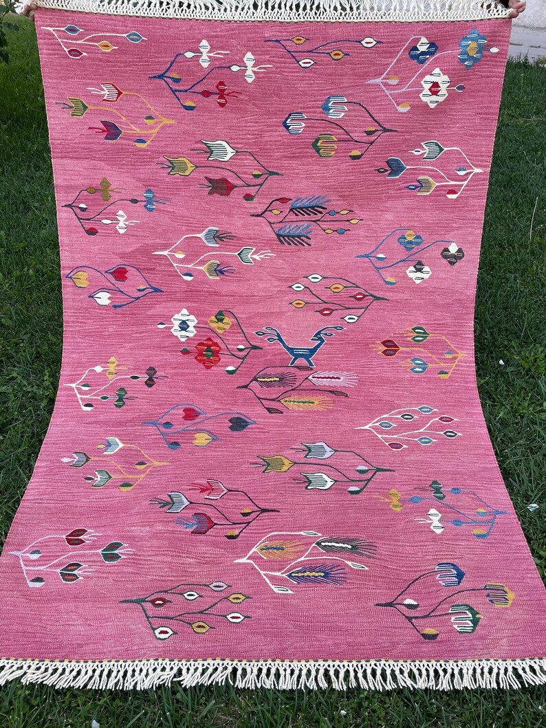May include: A pink rug with a geometric pattern of stylized flowers and leaves in shades of blue, green, yellow, and red. The rug has a white fringe border.