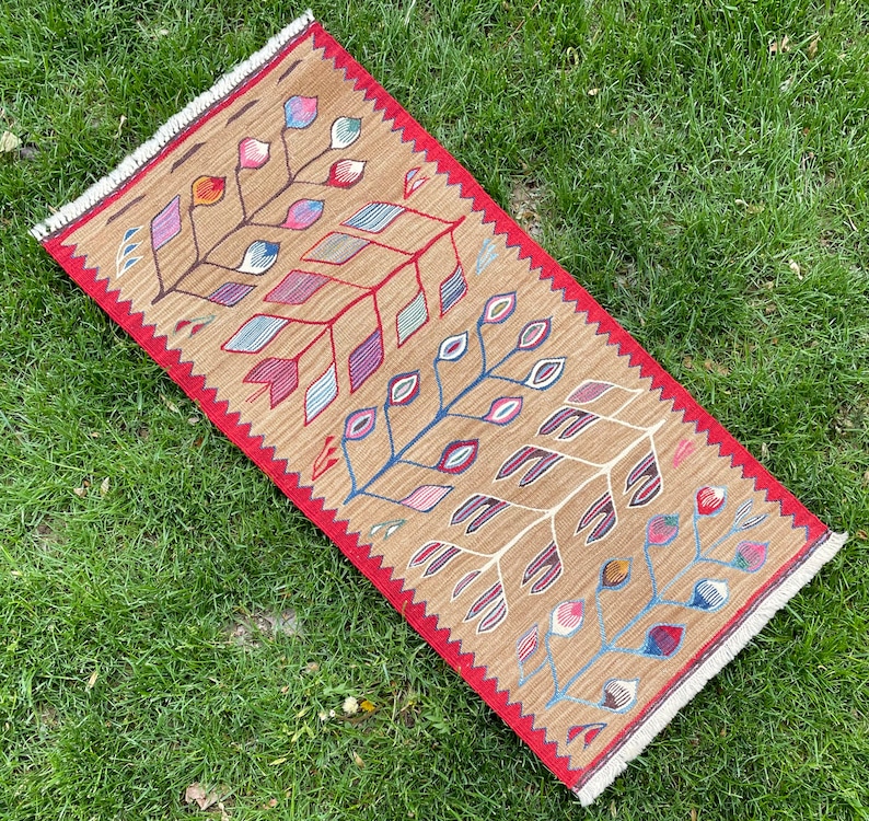 May include: A brown and red rug with a geometric pattern of stylized leaves and flowers. The rug has a fringed edge.