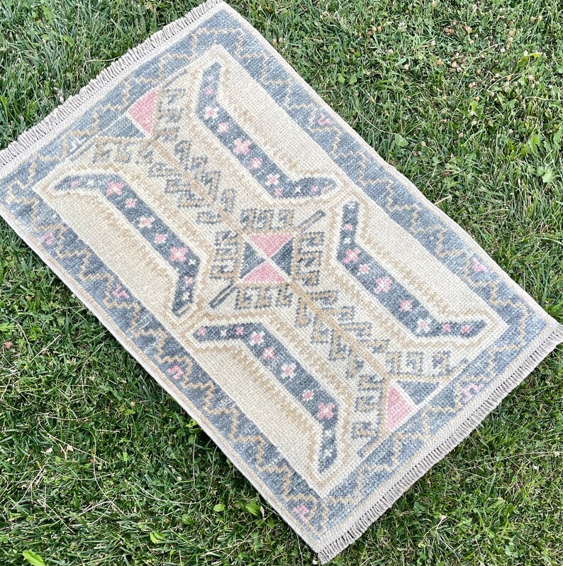 Puede incluir: Una alfombra beige con un patr&oacute;n geom&eacute;trico en tonos grises y rosas. La alfombra tiene un borde con flecos.
