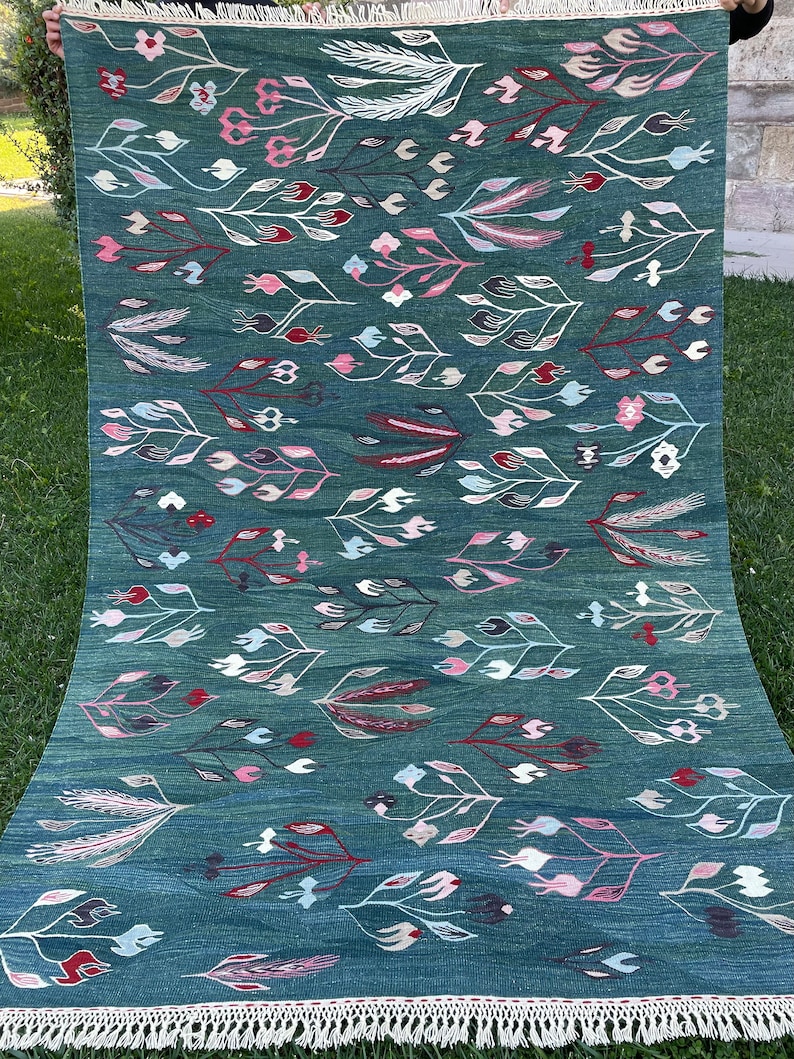 May include: A teal blue rug with a floral pattern. The rug has a white fringe border and features a repeating design of stylized flowers and leaves in shades of pink, white, and red.