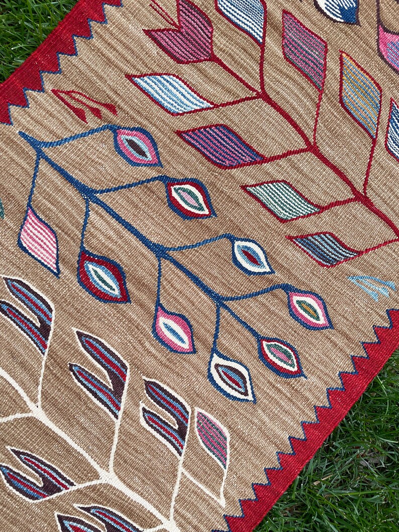May include: A brown and red patterned rug with a geometric design featuring stylized leaves and eyes. The rug has a fringed edge.