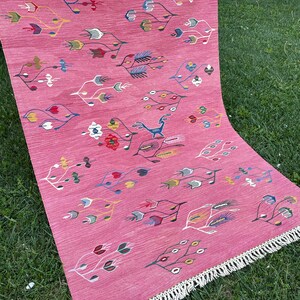 May include: A pink rug with a geometric pattern of stylized leaves and flowers in shades of blue, green, yellow, and red. The rug has a white fringe border.