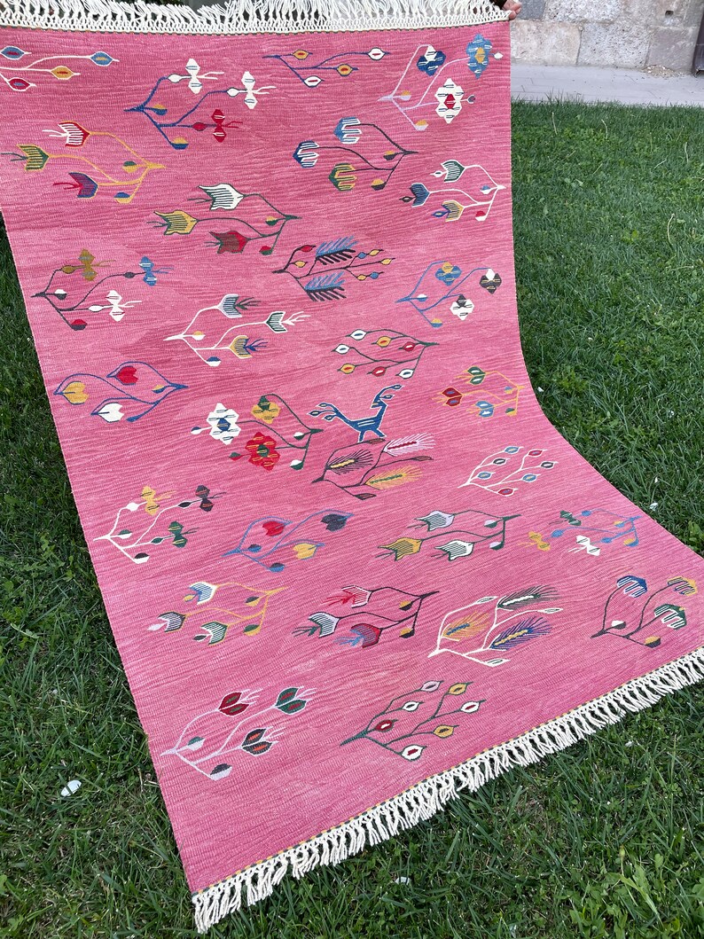 May include: A pink rug with a geometric pattern of stylized leaves and flowers in shades of blue, green, yellow, and red. The rug has a white fringe border.