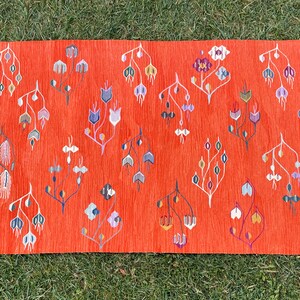 Pu&ograve; includere: Un tappeto rosso con un bordo di frange bianche presenta un motivo ripetitivo di disegni floreali stilizzati in tonalit&agrave; di blu, viola, verde e arancio.
