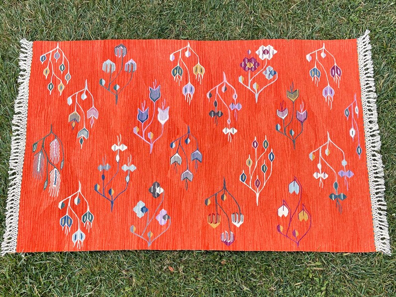 Pu&ograve; includere: Un tappeto rosso con un bordo di frange bianche presenta un motivo ripetitivo di disegni floreali stilizzati in tonalit&agrave; di blu, viola, verde e arancio.
