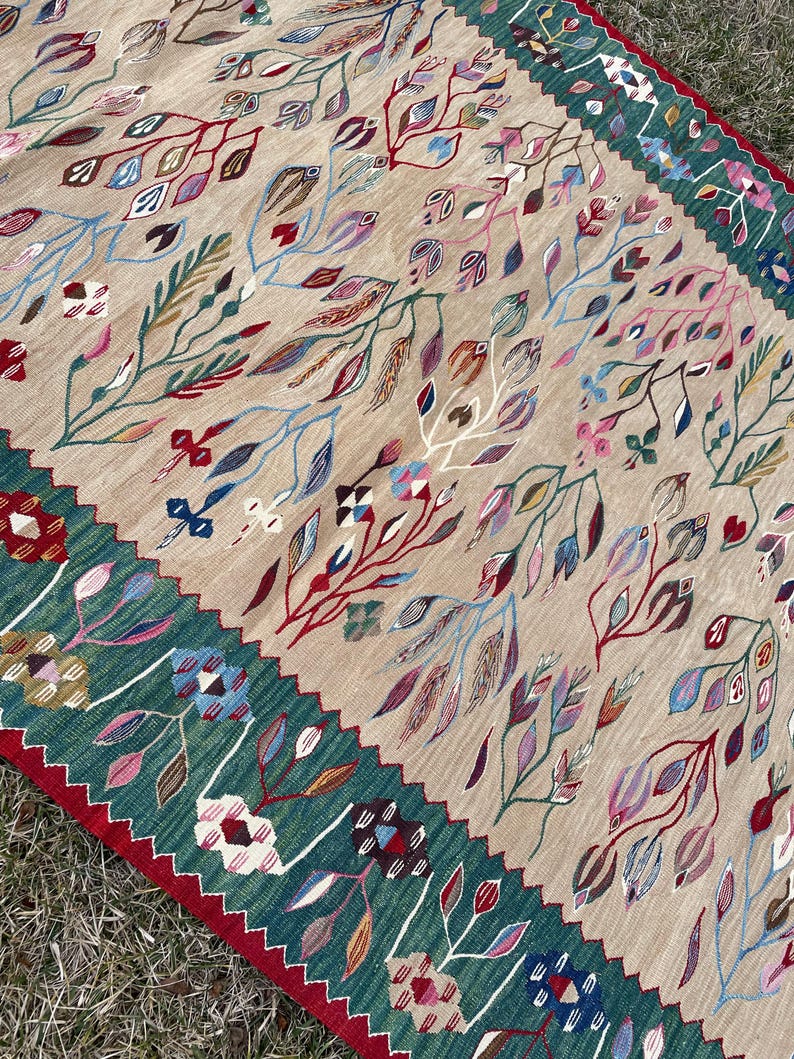 May include: A beige rug with a green border and red accents. The rug features a floral pattern with colourful leaves and flowers.