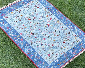 May include: A blue and red patterned rug with a floral design. The rug has a fringe border and is made of wool.