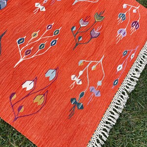 Pu&ograve; includere: Un tappeto rosso con un motivo geometrico di fiori e foglie stilizzati in tonalit&agrave; di viola, blu, verde, giallo e bianco. Il tappeto ha un bordo di frange bianche.