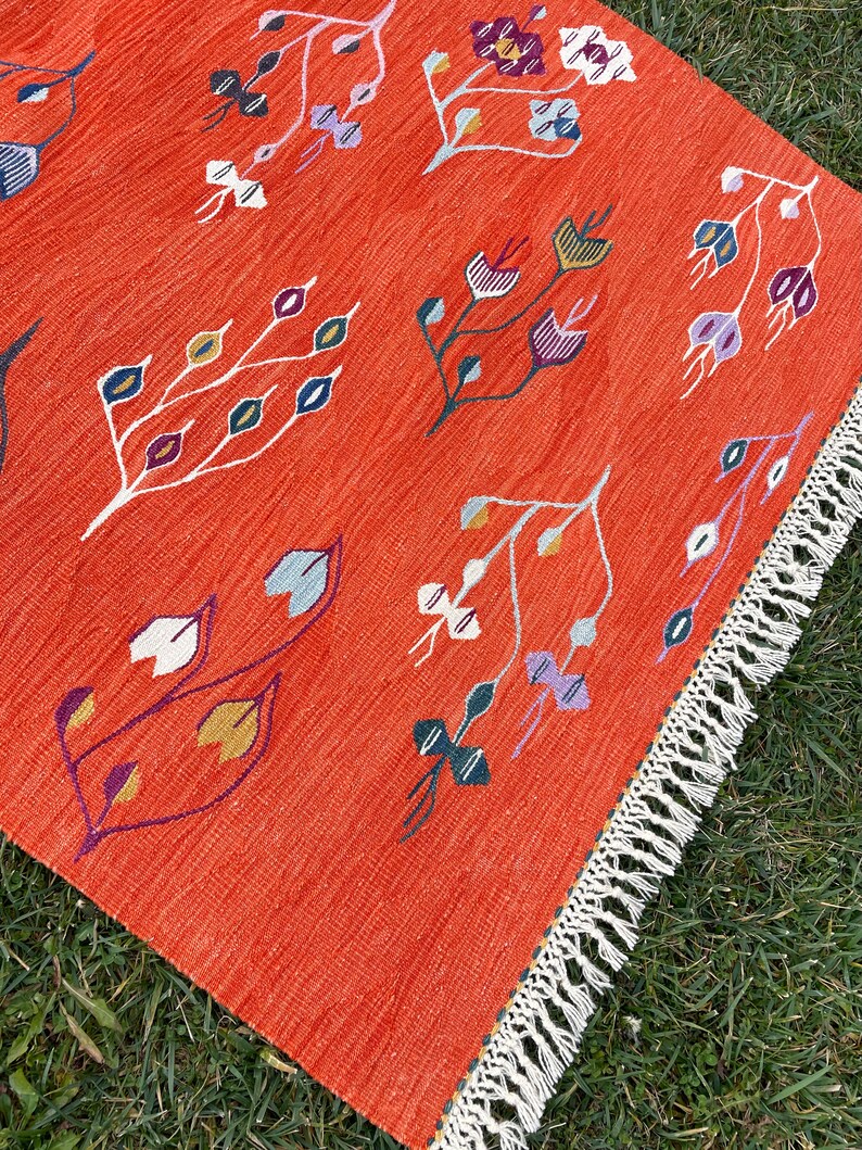 Pu&ograve; includere: Un tappeto rosso con un motivo geometrico di fiori e foglie stilizzati in tonalit&agrave; di viola, blu, verde, giallo e bianco. Il tappeto ha un bordo di frange bianche.
