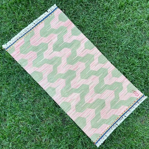 May include: A pink and green patterned rug with a fringed border. The rug has a geometric design with a zig-zag pattern.