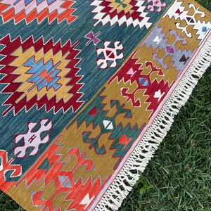 May include: A colourful rug with a geometric pattern in shades of red, blue, green, yellow, and white. The rug has a fringe border and is laid out on a green grassy surface.