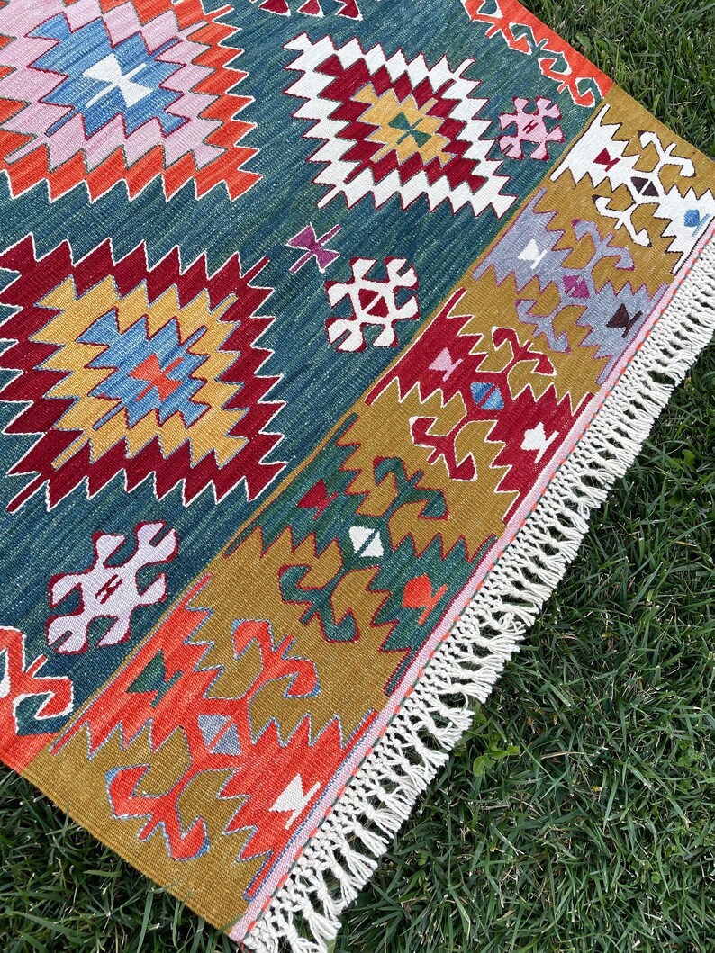 May include: A colourful rug with a geometric pattern in shades of red, blue, green, yellow, and white. The rug has a fringe border and is laid out on a green grassy surface.