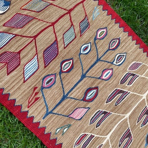 May include: A brown rug with a red border and a geometric pattern of stylized leaves and eyes in shades of blue, red, pink, and green.