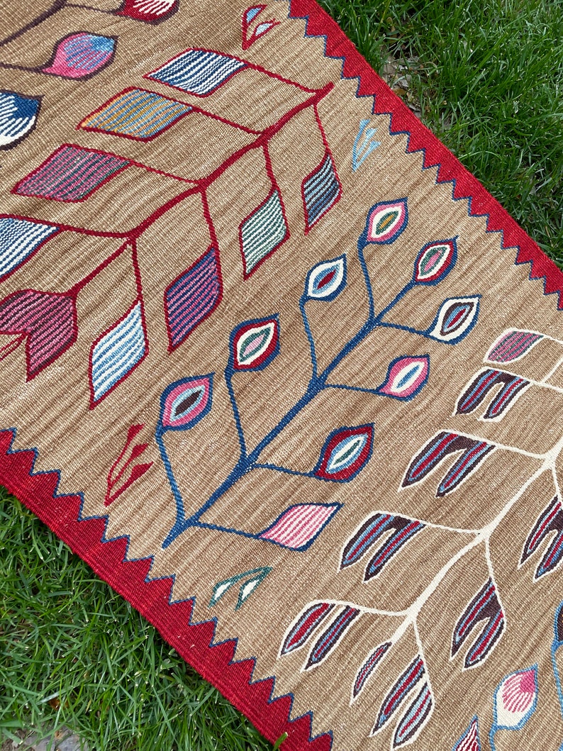 May include: A brown rug with a red border and a geometric pattern of stylized leaves and eyes in shades of blue, red, pink, and green.