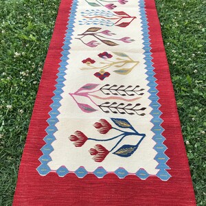 May include: A long, rectangular rug with a white background and a red border. The rug features a repeating pattern of stylized flowers in shades of red, blue, green, and brown.