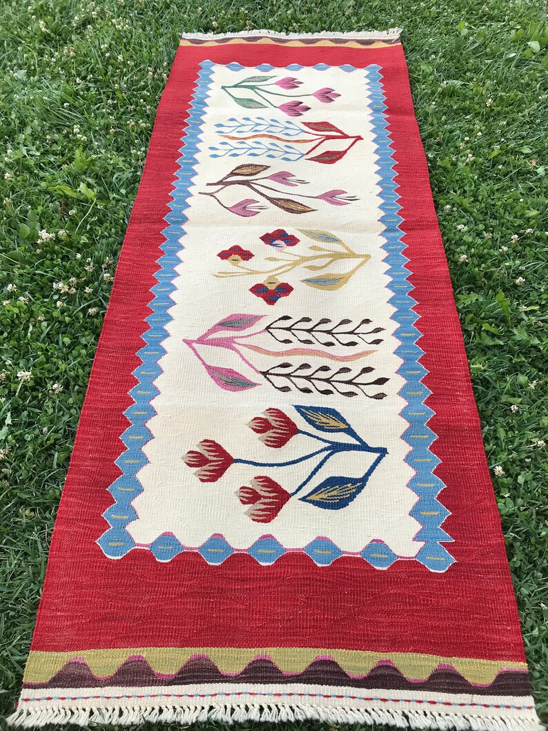 May include: A long, rectangular rug with a white background and a red border. The rug features a repeating pattern of stylized flowers in shades of red, blue, green, and brown.