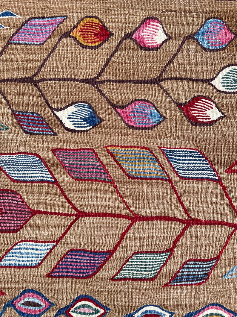 May include: A close-up of a woven fabric with a geometric pattern. The pattern features red, blue, pink, and yellow colors. The fabric has a brown background.