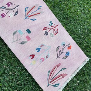 May include: A pink rug with a floral pattern. The rug has a fringe border and features stylized flowers in shades of red, blue, green, and yellow.