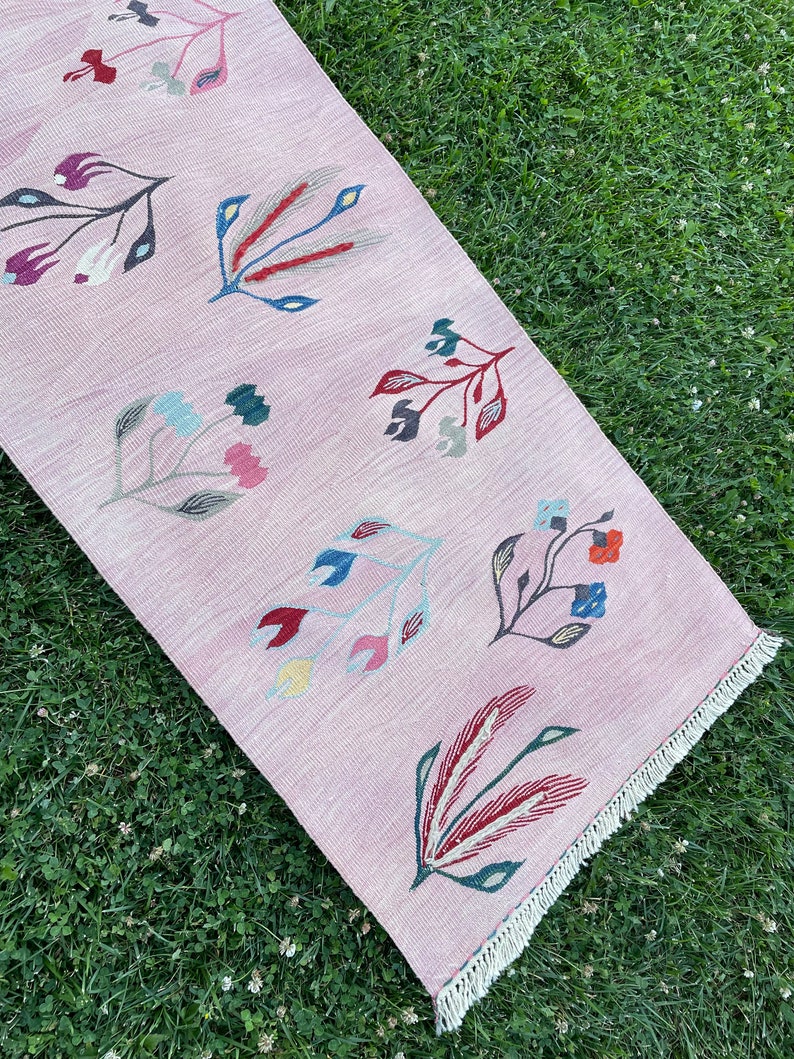 May include: A pink rug with a floral pattern. The rug has a fringe border and features stylized flowers in shades of red, blue, green, and yellow.