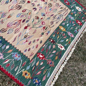 May include: A hand-woven rug with a floral pattern in shades of red, green, blue, and pink. The rug has a fringe border and is made of wool.