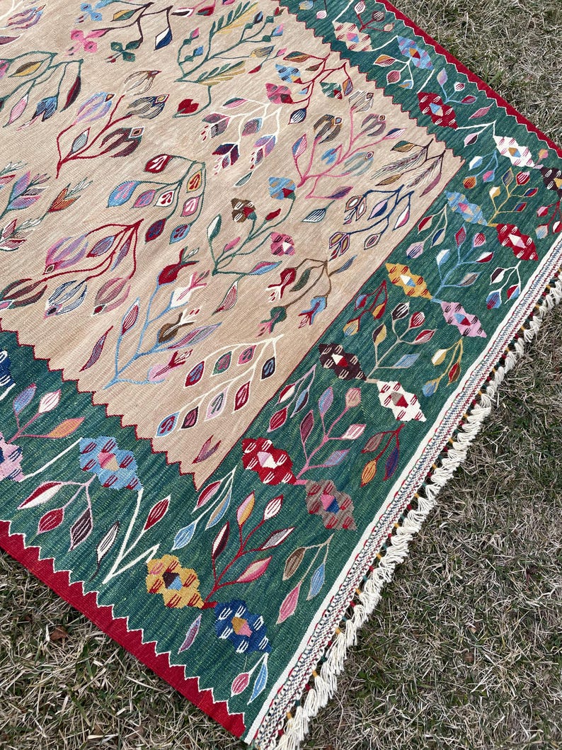 May include: A hand-woven rug with a floral pattern in shades of red, green, blue, and pink. The rug has a fringe border and is made of wool.