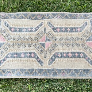 Puede incluir: Una alfombra peque&ntilde;a y rectangular con un patr&oacute;n geom&eacute;trico en tonos beige, gris y rosa. La alfombra tiene un borde con flecos.