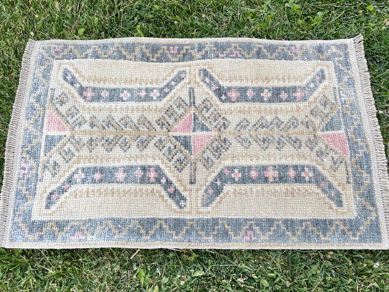 Puede incluir: Una alfombra peque&ntilde;a y rectangular con un patr&oacute;n geom&eacute;trico en tonos beige, gris y rosa. La alfombra tiene un borde con flecos.